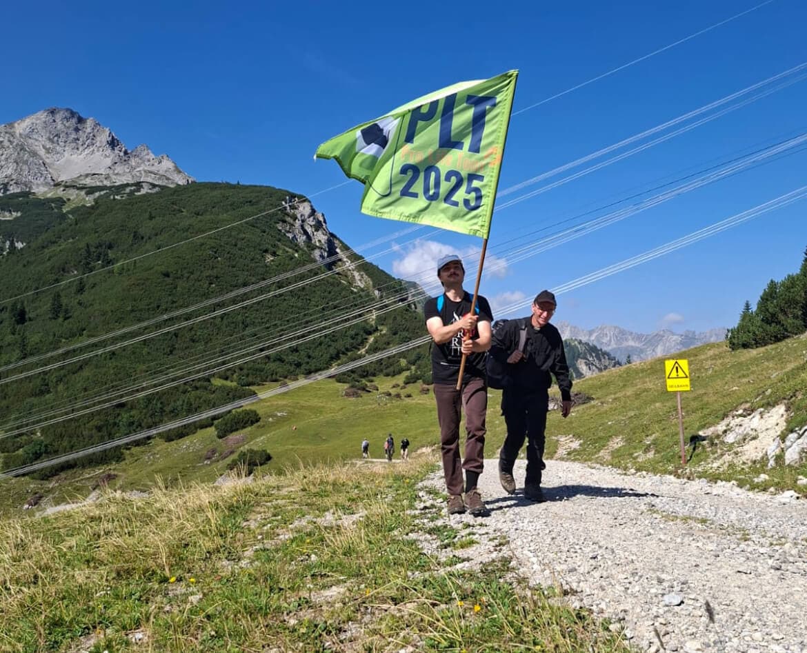 Zwei Teilnehmer der Pro Life Tour 2025 wandern mit der grünen „PLT 2025“-Fahne über einen Bergweg.
